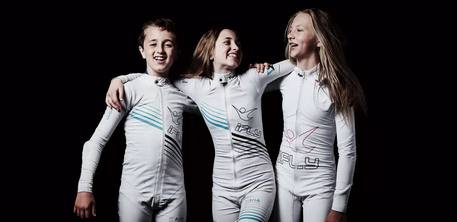 Three children wearing iFLY suits laughing with their arms around one another